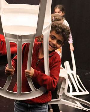 A boy laughs into the camera and holds up an upturned chair