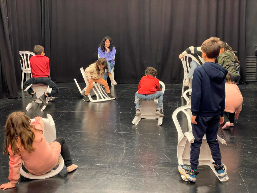 Eine Gruppe von Kindern mit ihrer Pädagogin im Theatersaal, die Kinder spielen mit Monobloc-Stühlen.
